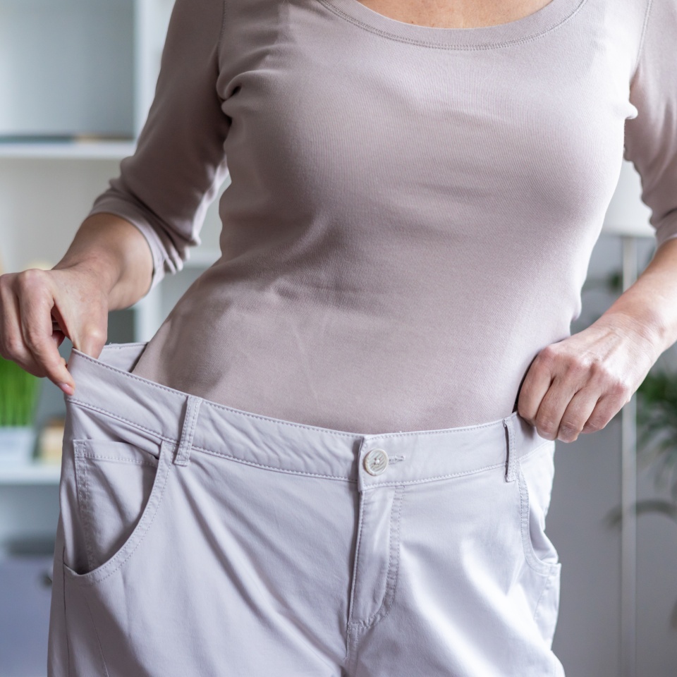 Woman showing weight loss achievement by holding oversized pants. Concept of fitness success, healthy lifestyle, diet, and personal transformation indoors.