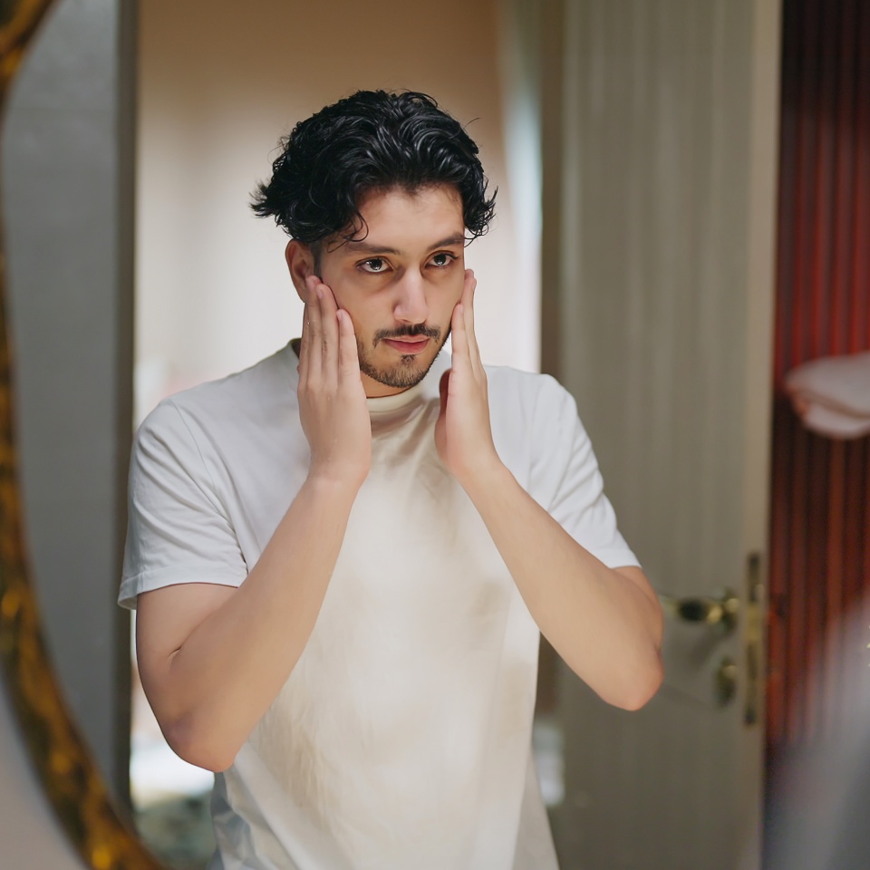 A man in a white t-shirt stands in front of a mirror, touching his face with both hands and looking at his reflection.