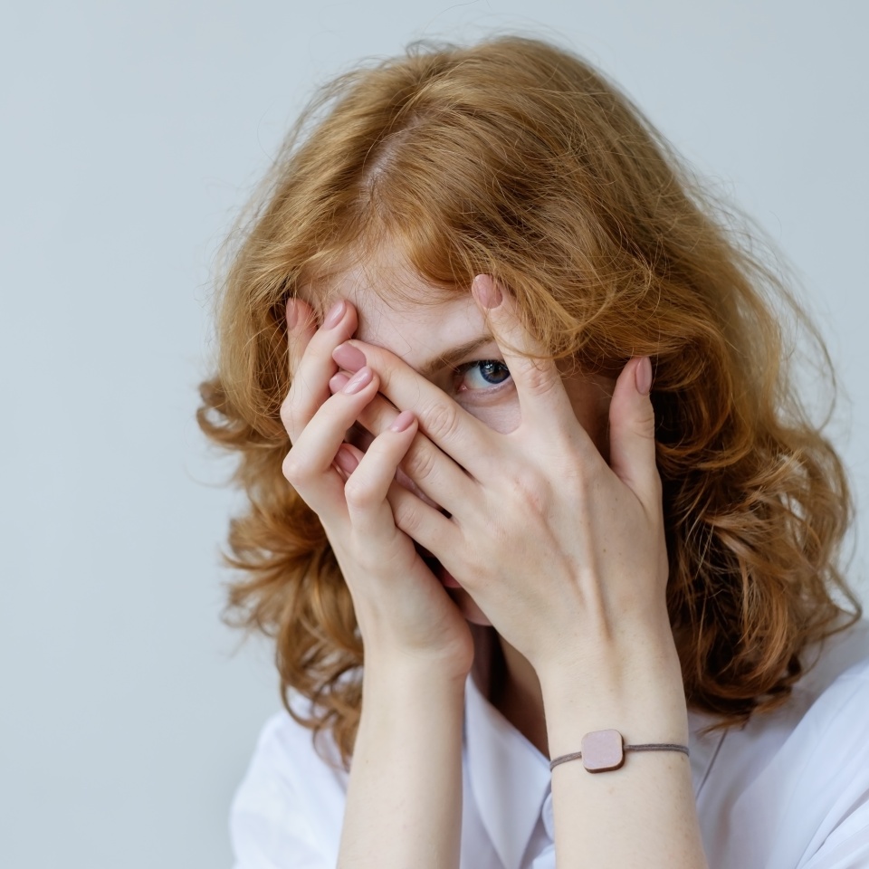A red-haired girl with curls in embarrassment covers her face with her hands and looks through her fingers. Social phobia of the younger generation.