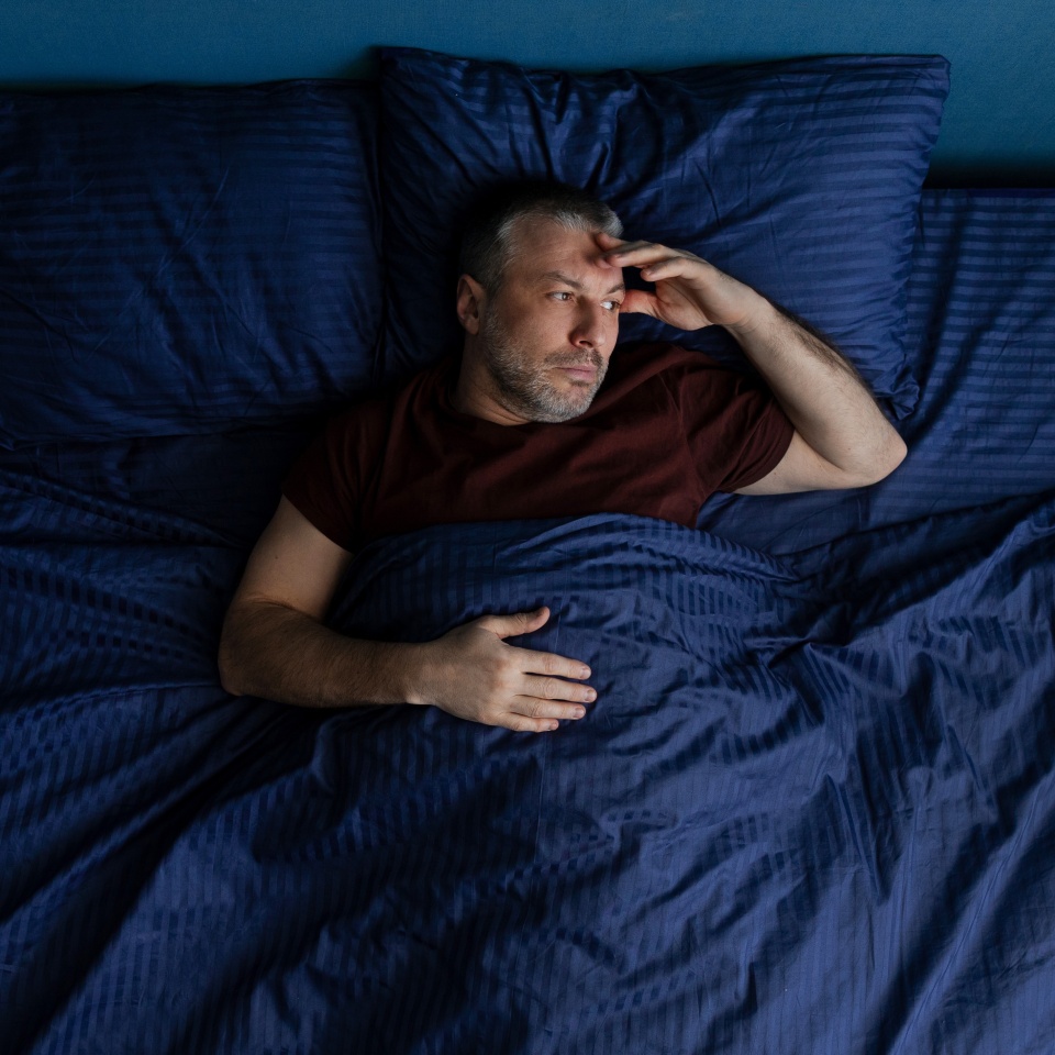 Top view of middle aged man lying in bed holding head with hand, worried expression, insomnia or stress concept, dark blue bedding in modern bedroom setting