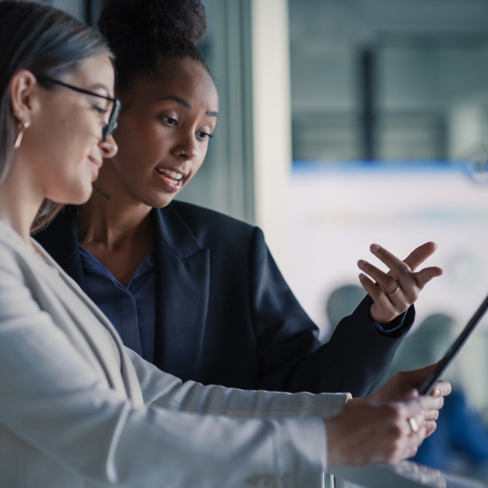 Two women in business attire stand indoors, discussing information displayed on a digital tablet.