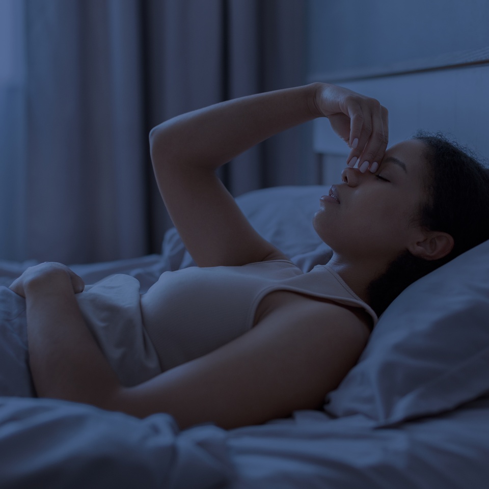 Black woman lying awake in bed at night and pinching her nose, suggesting sinus pressure, headache, insomnia and discomfort in a dark blue bedroom