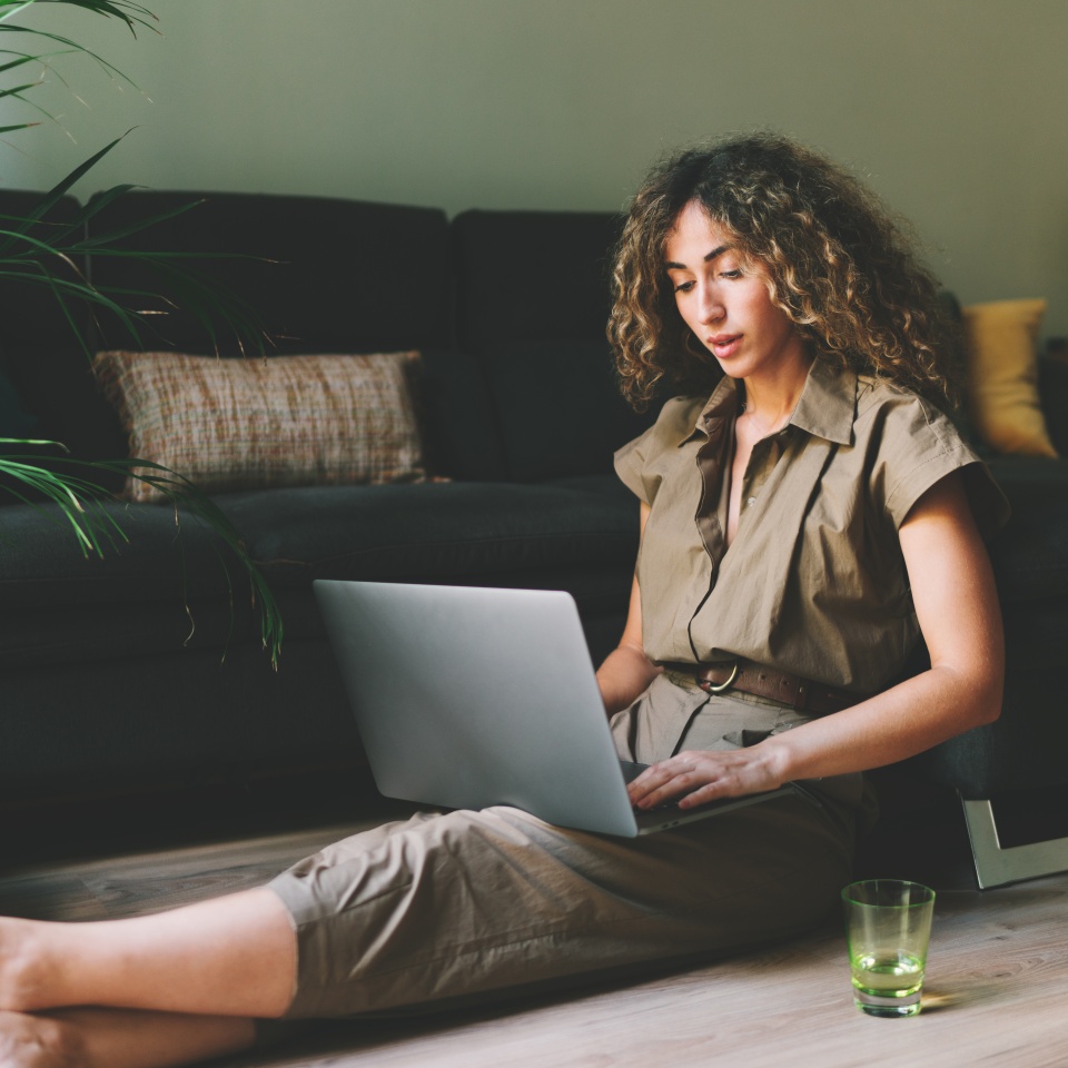 Beautiful female in casual outfit chilling on a floor beside comfortable couch in a living room with laptop on her knees. Web designer browsing laptop while working on a remote project at home.