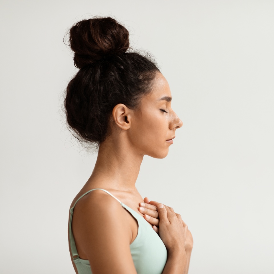Profile portrait of calm young woman with hands on chest and closed eyes, side view shot of peaceful millennial female meditating or praying while standing over white background indoors, copy space