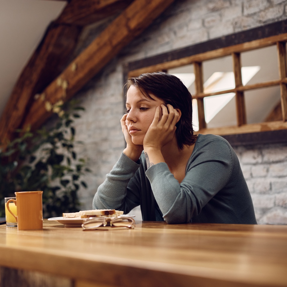 A woman sits at a wooden table with her head in her hands, looking at a plate with toast and a mug, appearing thoughtful or tired.