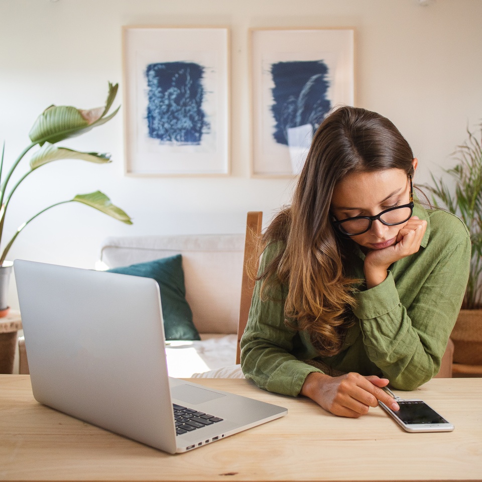 Woman sitting at a desk with a laptop, looking at her smartphone. She wears glasses and a green top. Two abstract artworks hang on the wall behind her.