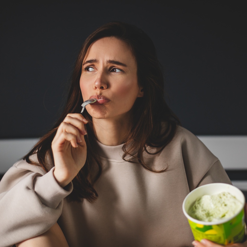 Woman in a beige sweatshirt holds a spoon to her lips and a tub of ice cream, looking thoughtfully to the side.