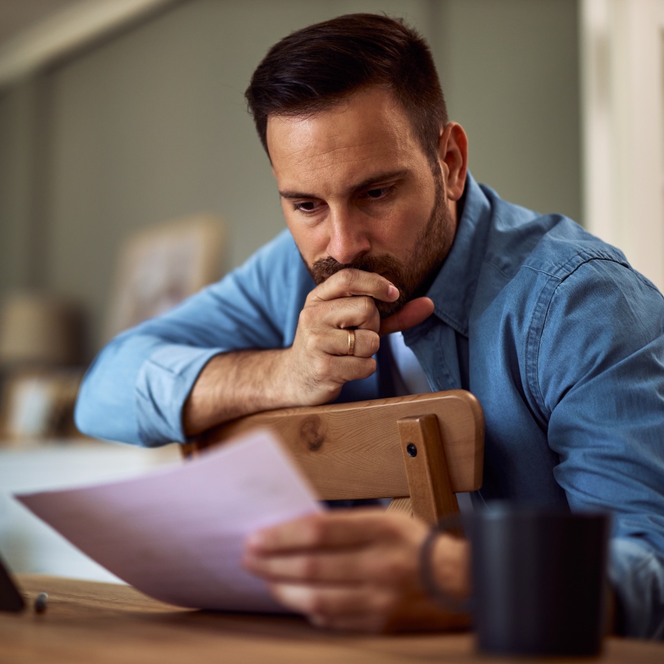 A focused, preoccupied young adult male freelancer reading and analyzing the documents in his hand with a hand over his mouth and sitting backwards in a chair in front of a tablet.