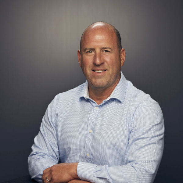 A man with a shaved head wearing a light blue button-down shirt sits at a table, smiling, against a plain dark background.