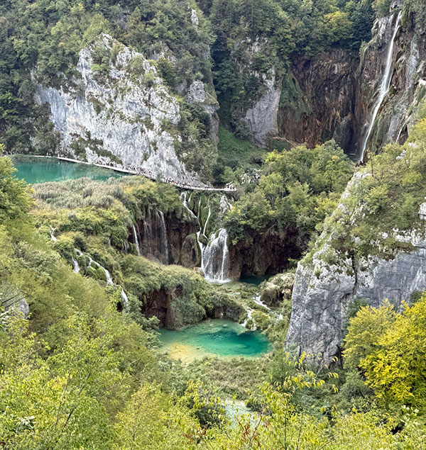 A scenic landscape with turquoise lakes, waterfalls, rocky cliffs, and dense green vegetation under a cloudy sky. A wooden footbridge crosses the water.