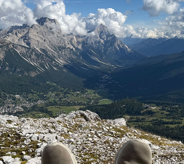 A person’s shoes are visible in the foreground with a rocky slope, forested valley, villages, and tall mountains under a partly cloudy sky in the background.
