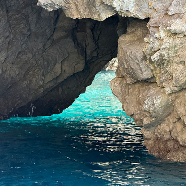 A rocky sea cave opening overlooks clear turquoise water, with sunlight illuminating part of the cave walls and the ocean below.