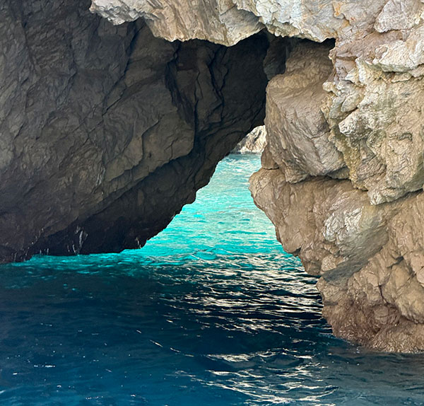 A rocky cave opening faces clear turquoise water, with sunlight illuminating the sea and the stone surfaces around the entrance.