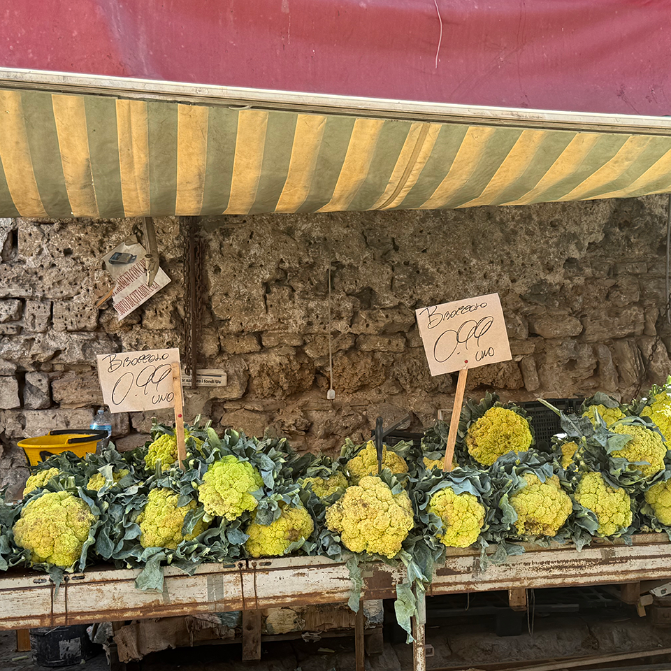 Yellow cauliflower heads with green leaves are displayed on a wooden stall under a striped canopy, with handwritten price signs reading “Broccolo” and “cuo”.